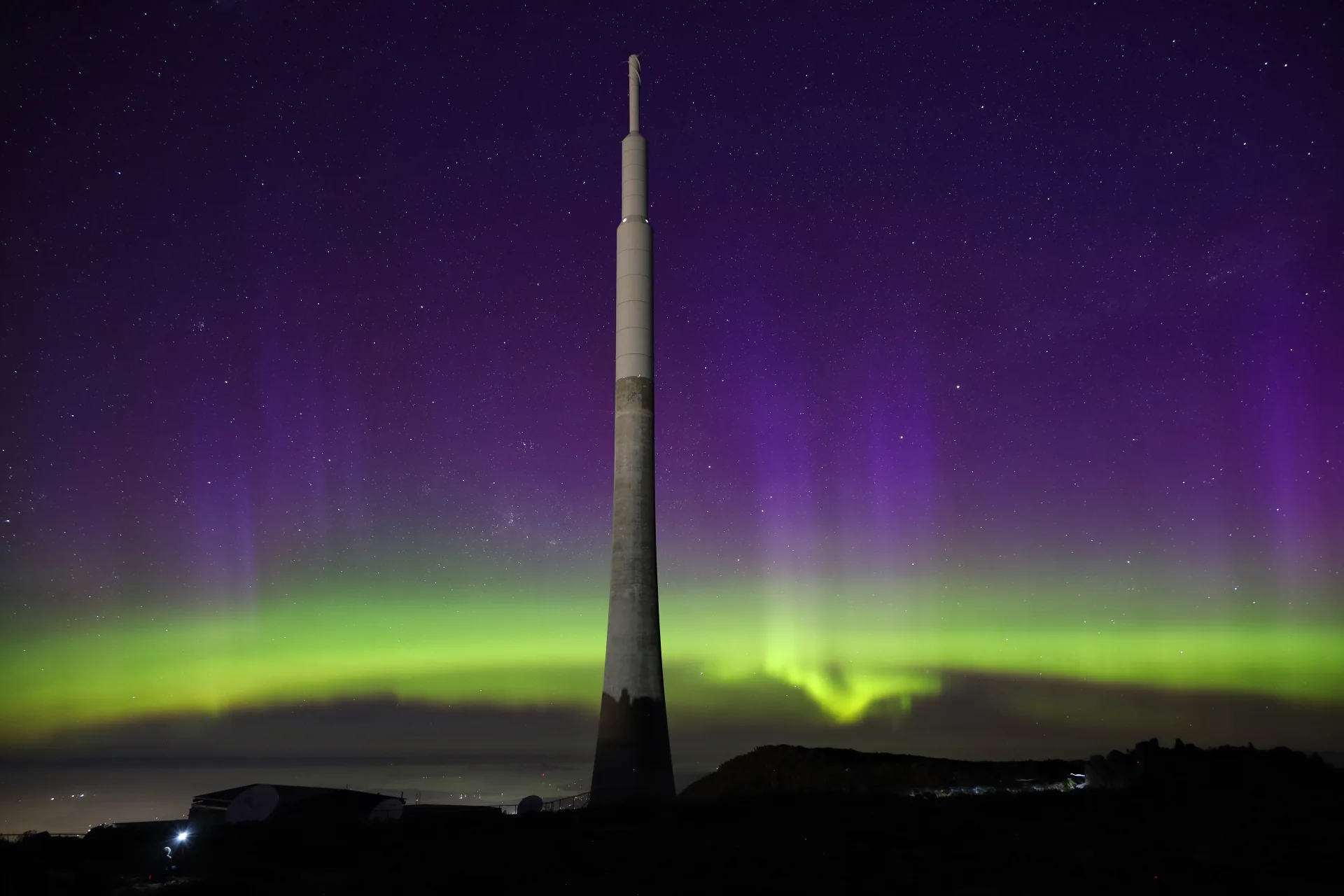 A photograph by Maddie of a night scene atop Mount Wellington / kunanyi, featuring a tall communications tower silhouetted against a vivid aurora australis. Bands of green and purple light stretch across a star-filled sky, creating a dramatic contrast with the dark foreground.