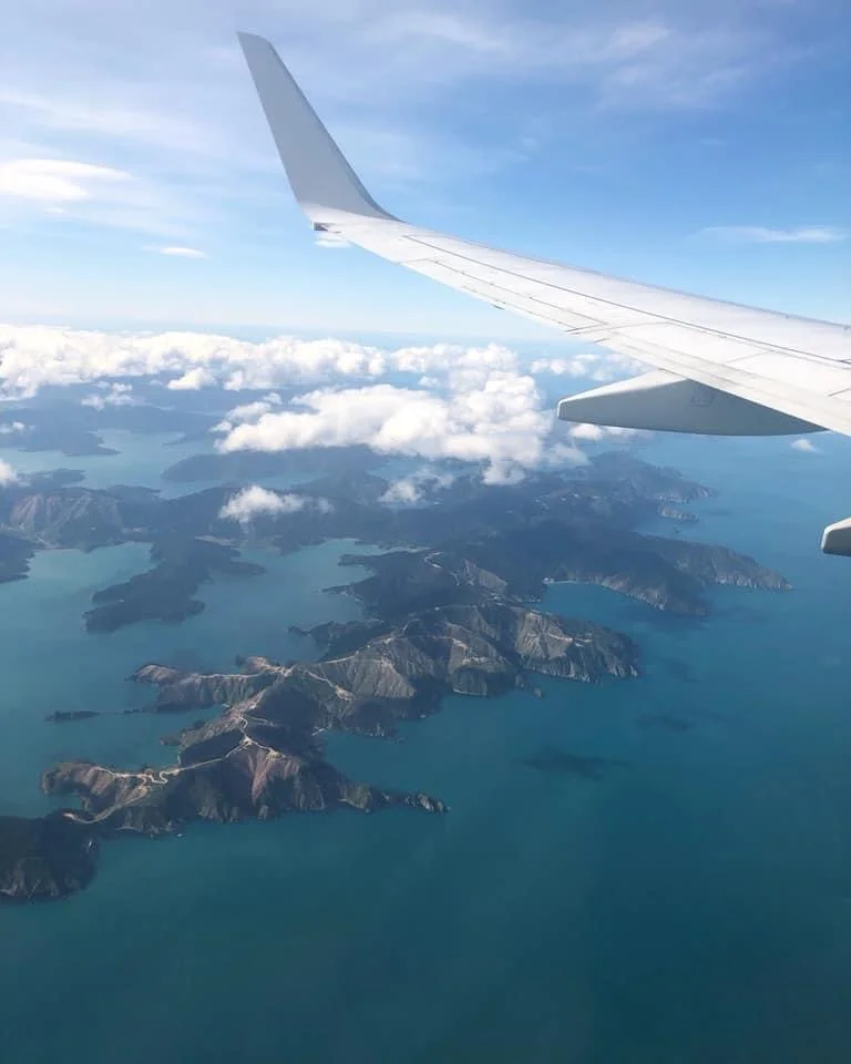 An aerial view from an airplane window, with the wing extending over a scattered archipelago of rugged islands surrounded by deep blue water. Light clouds float above, partially veiling the landscape below.