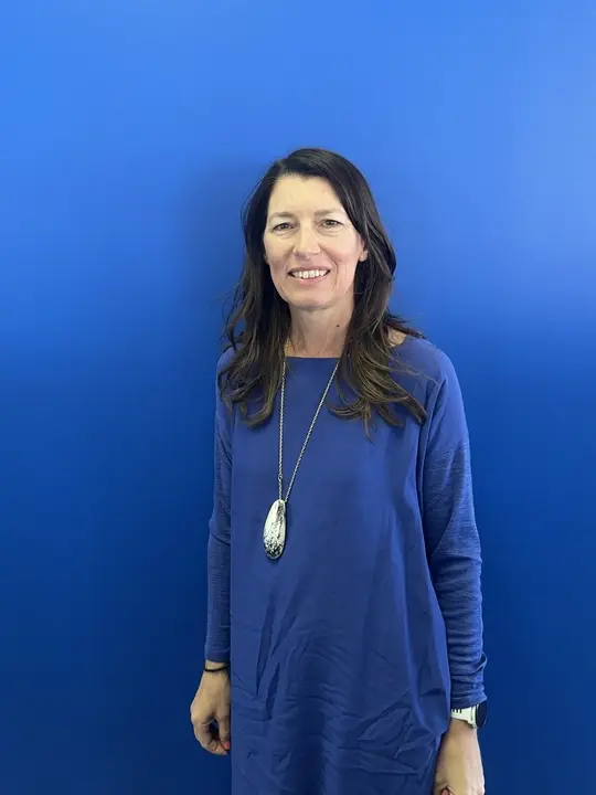 A professional headshot of Narelle Walch against a blue background. She wears a blue long sleeve dress, with Dinosaur Designs pendant and is smiling directly at the camera.