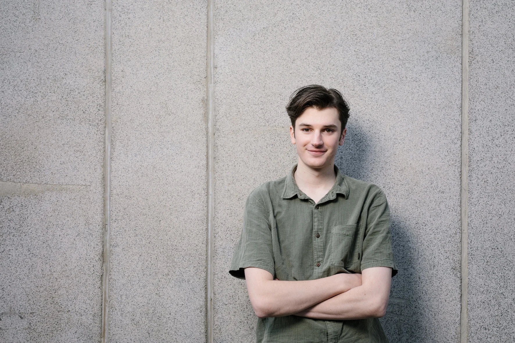 A portrait of Ned Daniels standing against a textured concrete wall, arms crossed and smiling calmly while looking at the camera. He wears a short-sleeved olive-green button-up linen shirt, his dark brown hair parted neatly to the side.