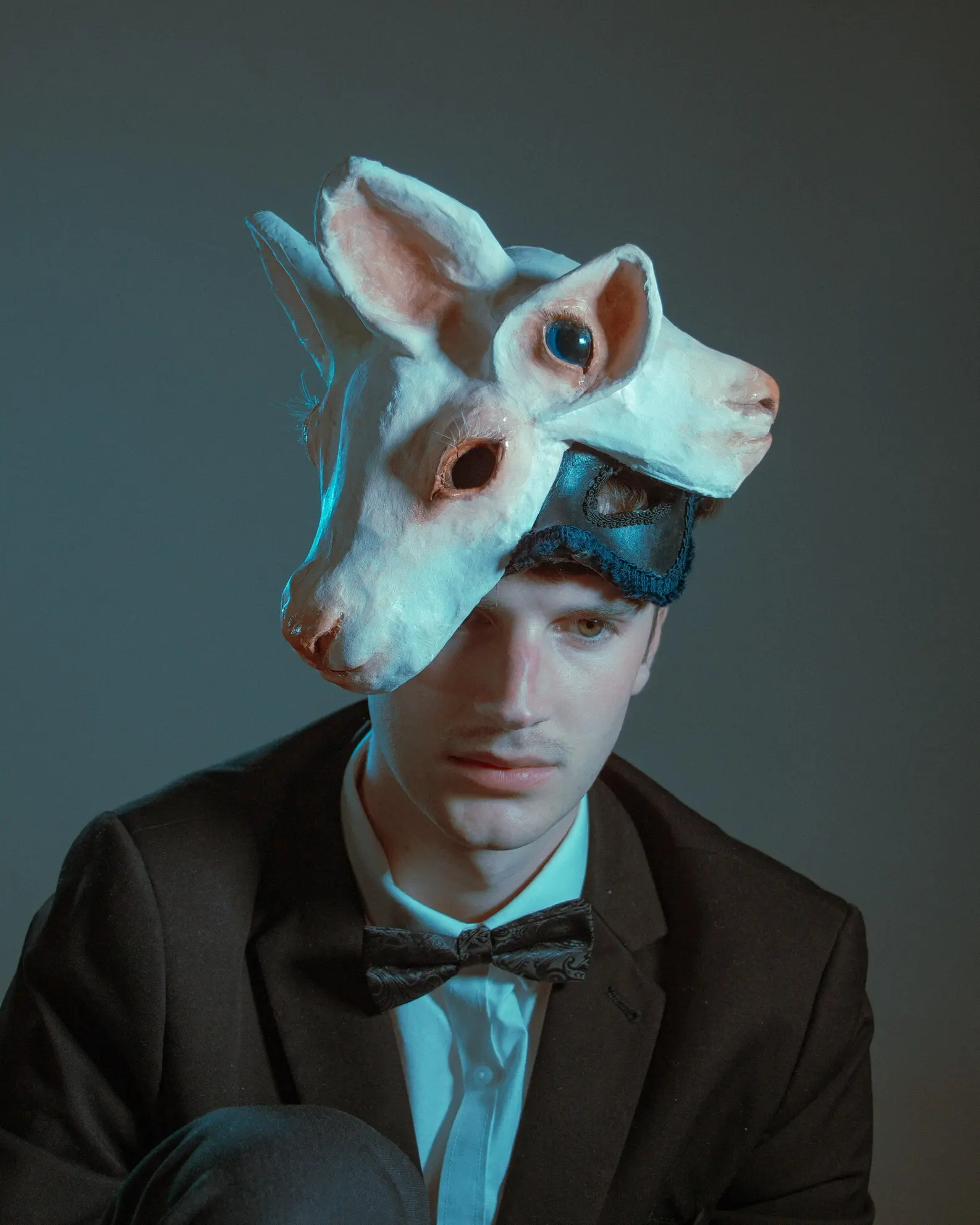 A stylised studio photograph of Ned wearing a sculptural conjoined lamb mask, created by himself for Dark Mofo 2023’s Blue Rose Ball. The mask features two fused lamb heads with hollowed eye sockets and delicate surface detailing, worn with a black suit and bow tie under dramatic, moody lighting. Materials: papier-mâché, cardboard, acrylic paint, resin, felt, horse hair. Inspired by Laura Gilpin’s poem “The Two-Headed Calf.”