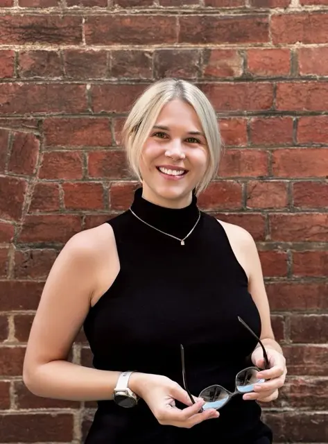 A portrait of Olivia Potter standing in front of a red brick wall, smiling warmly at the camera. She wears a sleeveless black high-neck top and a delicate necklace, with her blonde hair cut in a short bob.