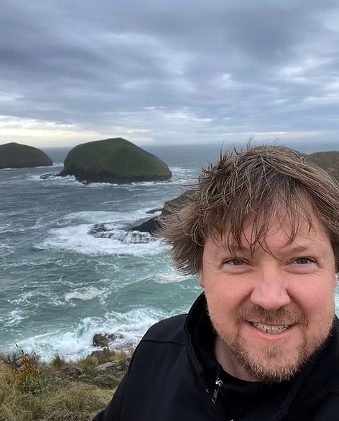 A smiling selfie of Pete on the top of a cliff at Cape Grim in North West Tasmania, with the churing ocean and a cloudy sky in the background.