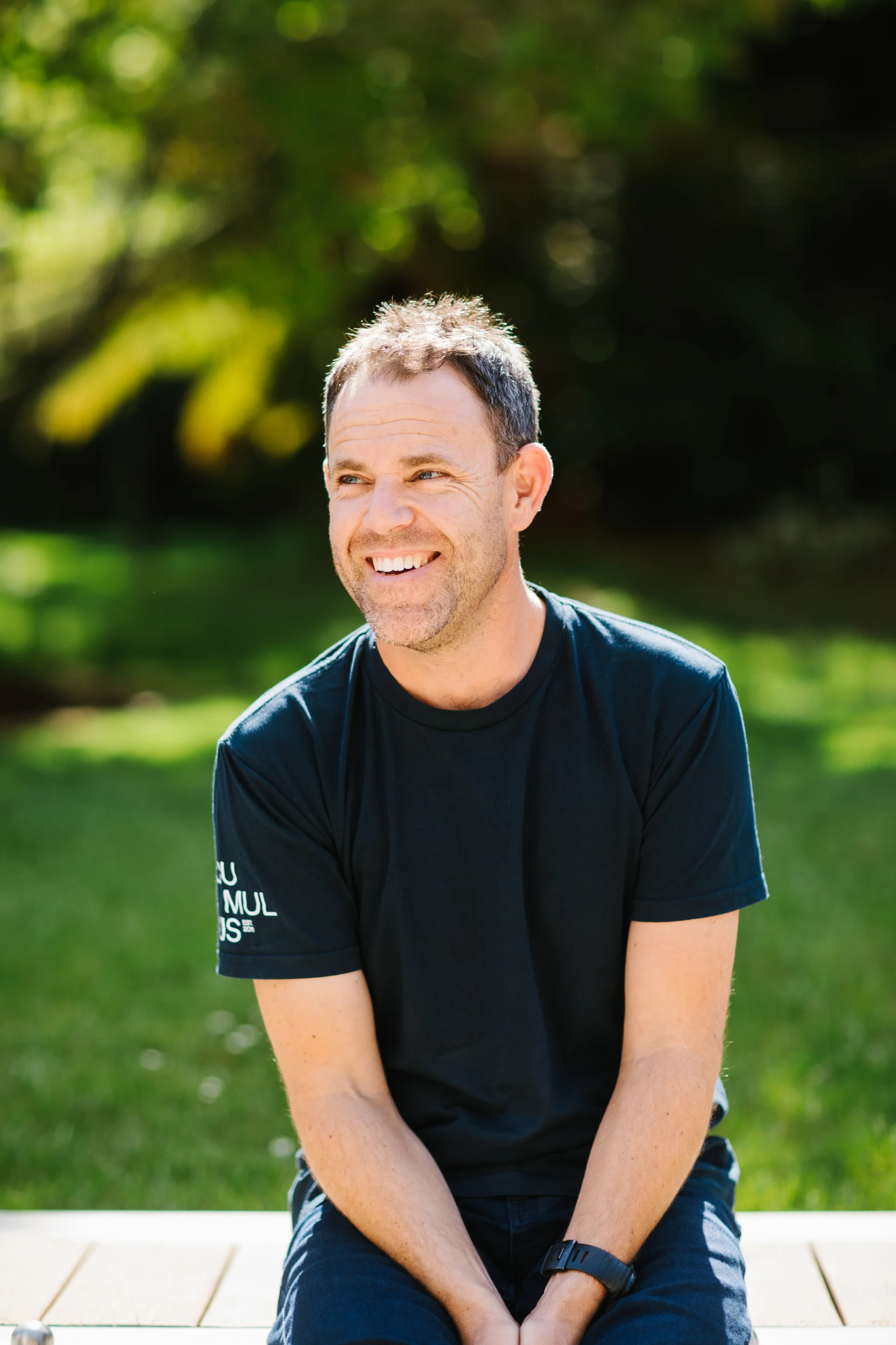 A professional portrait of Phil Ackerly sitting outdoors in bright natural light, smiling and looking off to the side. He wears a black t-shirt, with a soft, blurred garden setting behind.