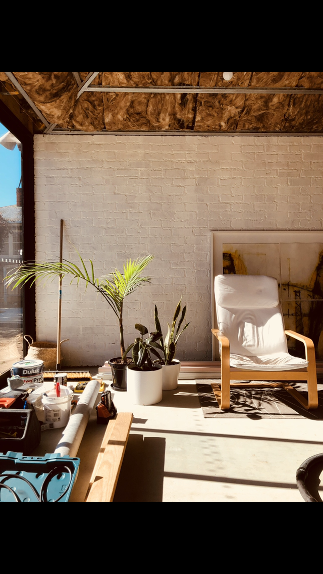A sunlit interior of a partially renovated space, with tools and materials in the foreground. A white lounge chair and potted plants sit against a painted brick wall while insulation in the ceiling is exposed.