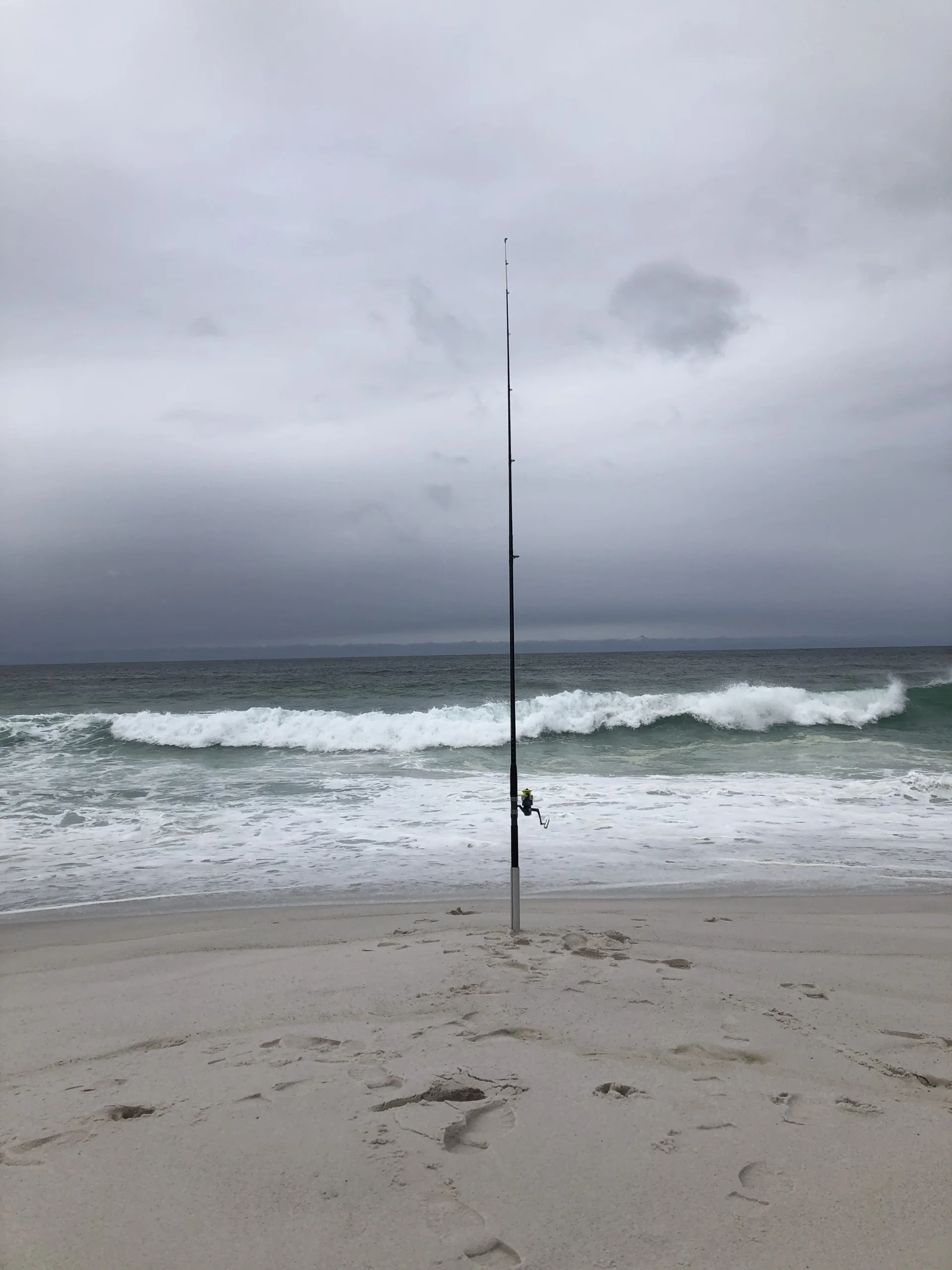 A minimal beach scene with a single fishing rod planted upright in the sand, facing the ocean. Small waves roll in beneath a grey, overcast sky, creating a quiet, solitary atmosphere.