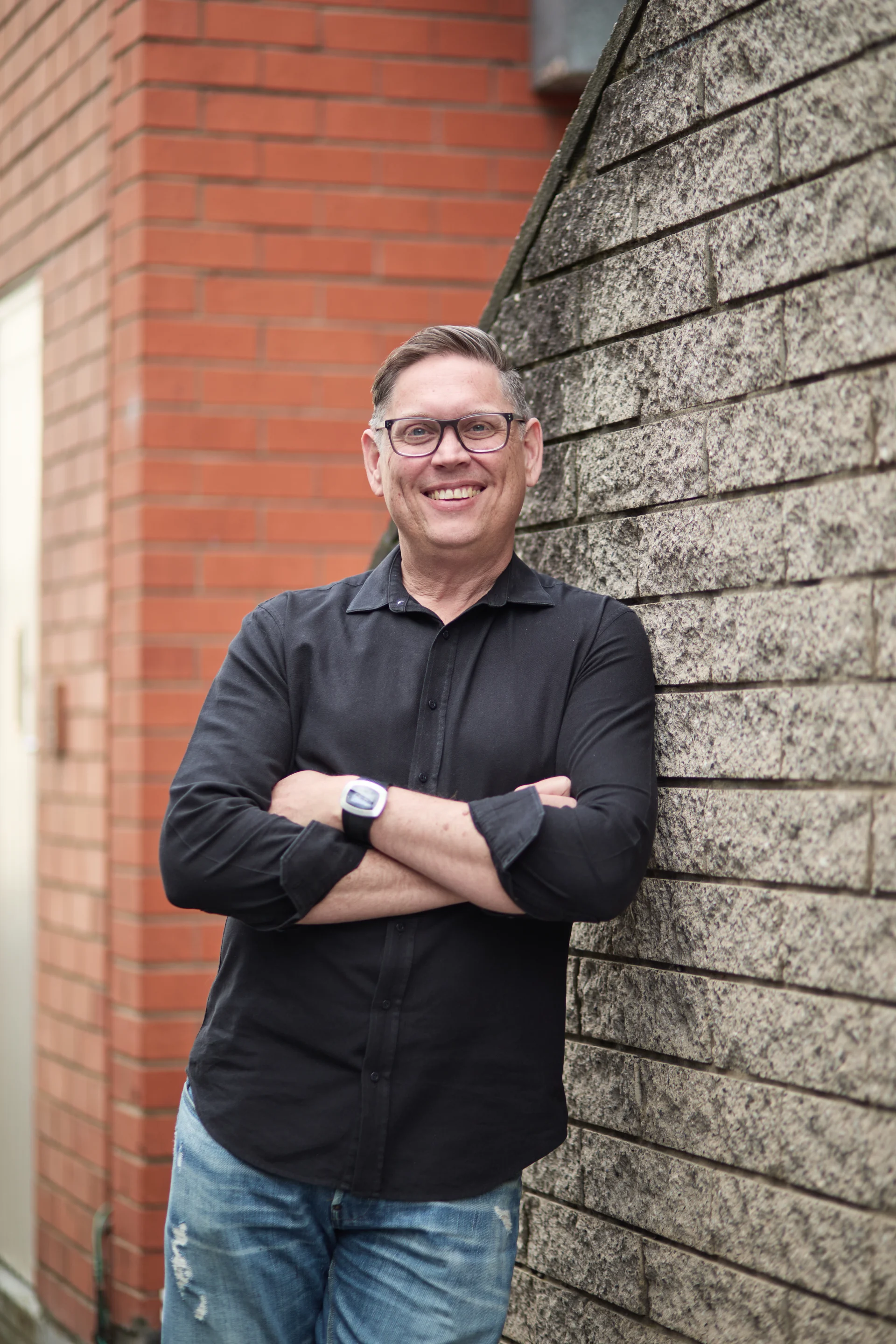 A professional headshot of Reuben Bourke standing outdoors leaning against a textured stone wall. He wears a black button-up shirt and glasses, arms folded, smiling directly at the camera with a friendly and confident expression.