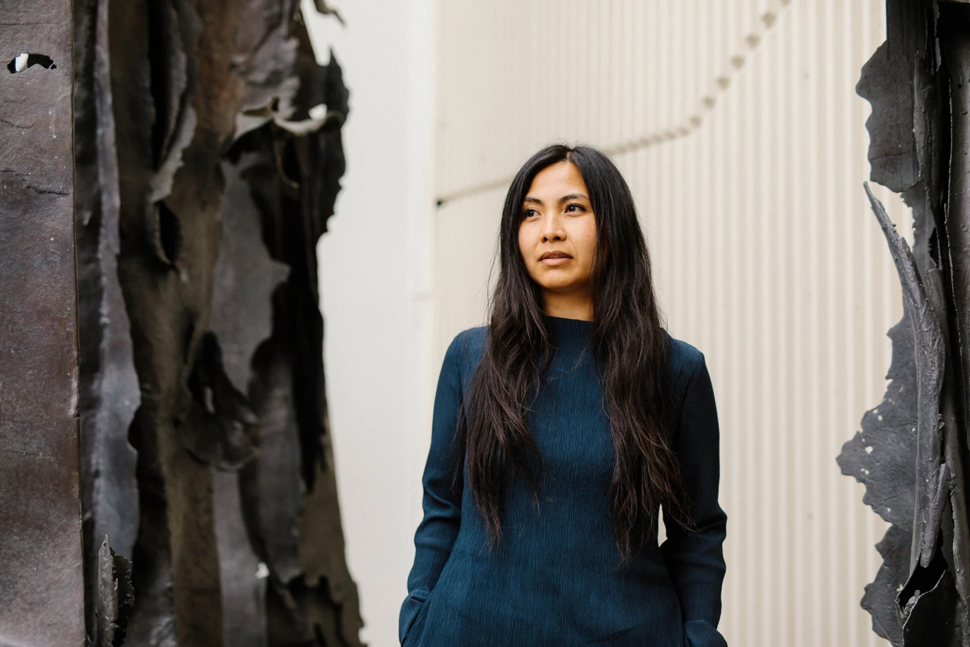 A professional headshot of Rosalyn Bermudez standing against a light, textured backdrop with bronze sculptures. She wears a dark blue long-sleeve top, her long dark hair worn loose, looking slightly off-camera with a calm and confident expression.