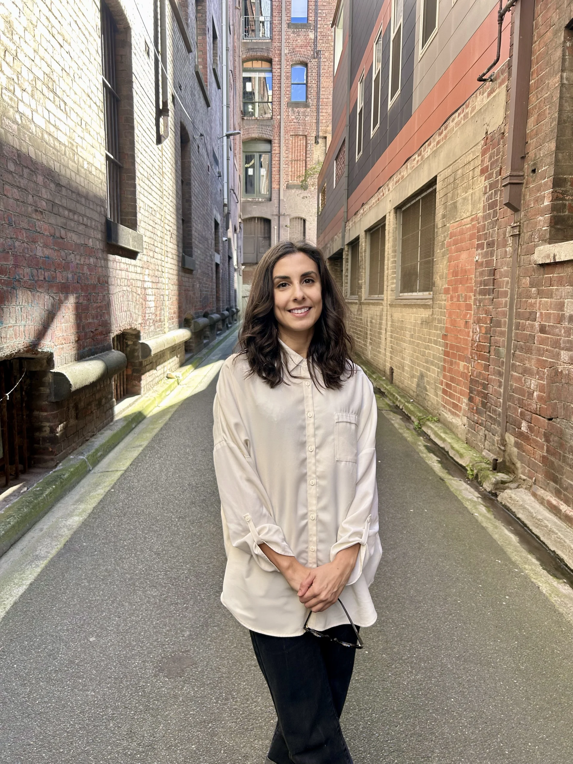 A professional portrait of Rosella Sciurti standing in a narrow urban laneway lined with brick buildings. She smiles at the camera, wearing a light long sleeve shirt, with soft daylight filtering into the space.