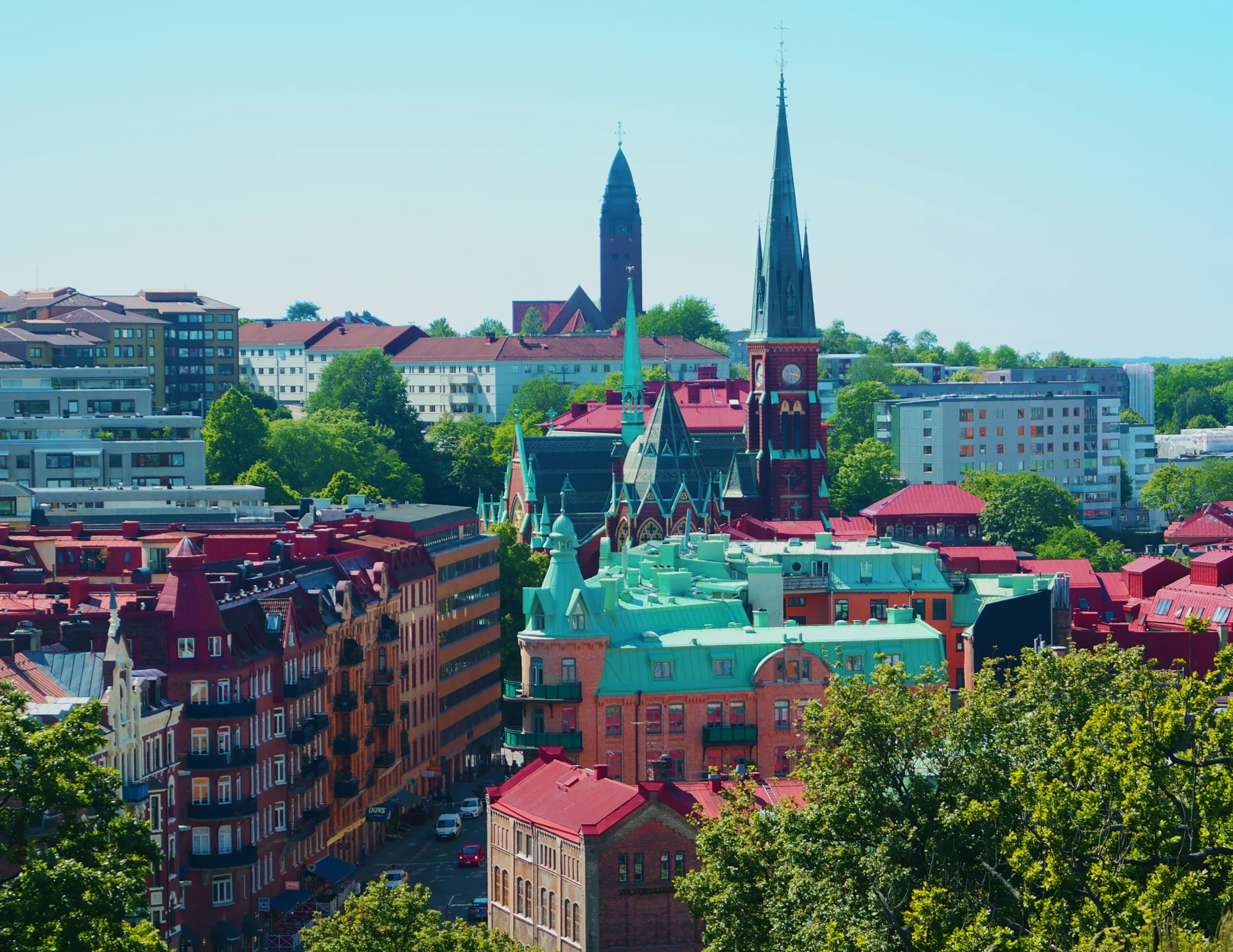 A vibrant cityscape featuring colourful rooftops and historic buildings with spires and towers rising above the urban fabric. The scene is lush with greenery and bathed in bright daylight.