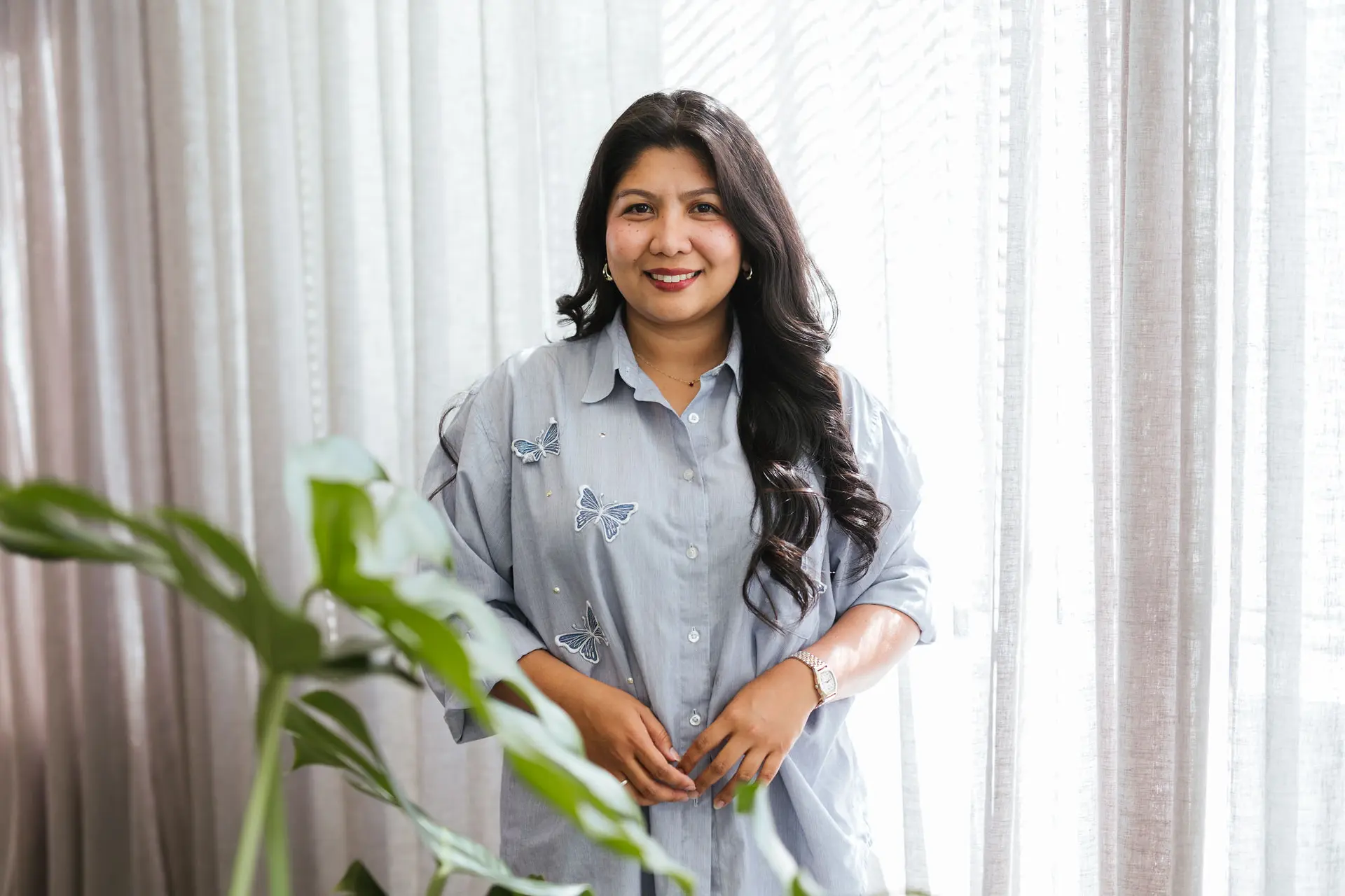 A professional portrait of Salve Reina Oarde standing beside a softly lit window, smiling warmly at the camera. She wears a light blue shirt with subtle embroidered butterfly details, with sheer curtains and indoor plants creating a calm, natural backdrop.