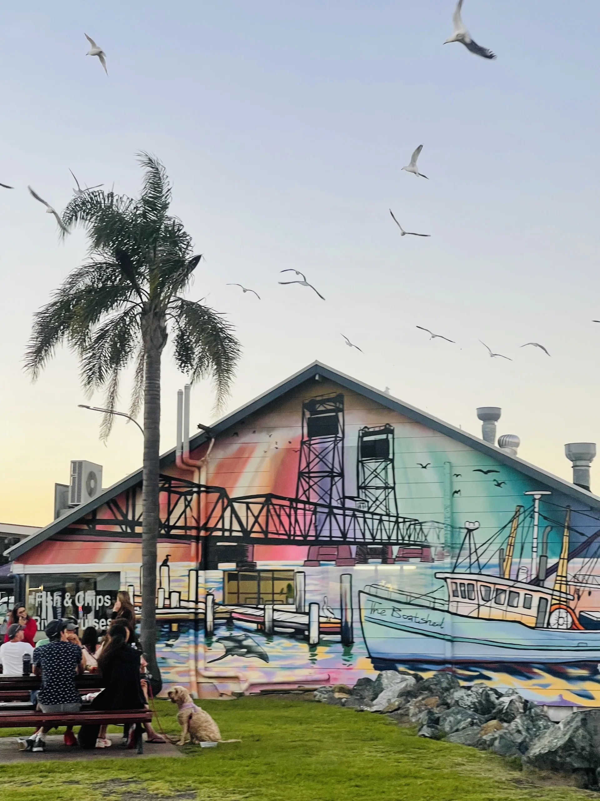A lively outdoor scene of a waterfront building covered in a colourful mural depicting boats and industrial structures. People gather at picnic tables in the foreground, while seagulls fly overhead against a soft evening sky.