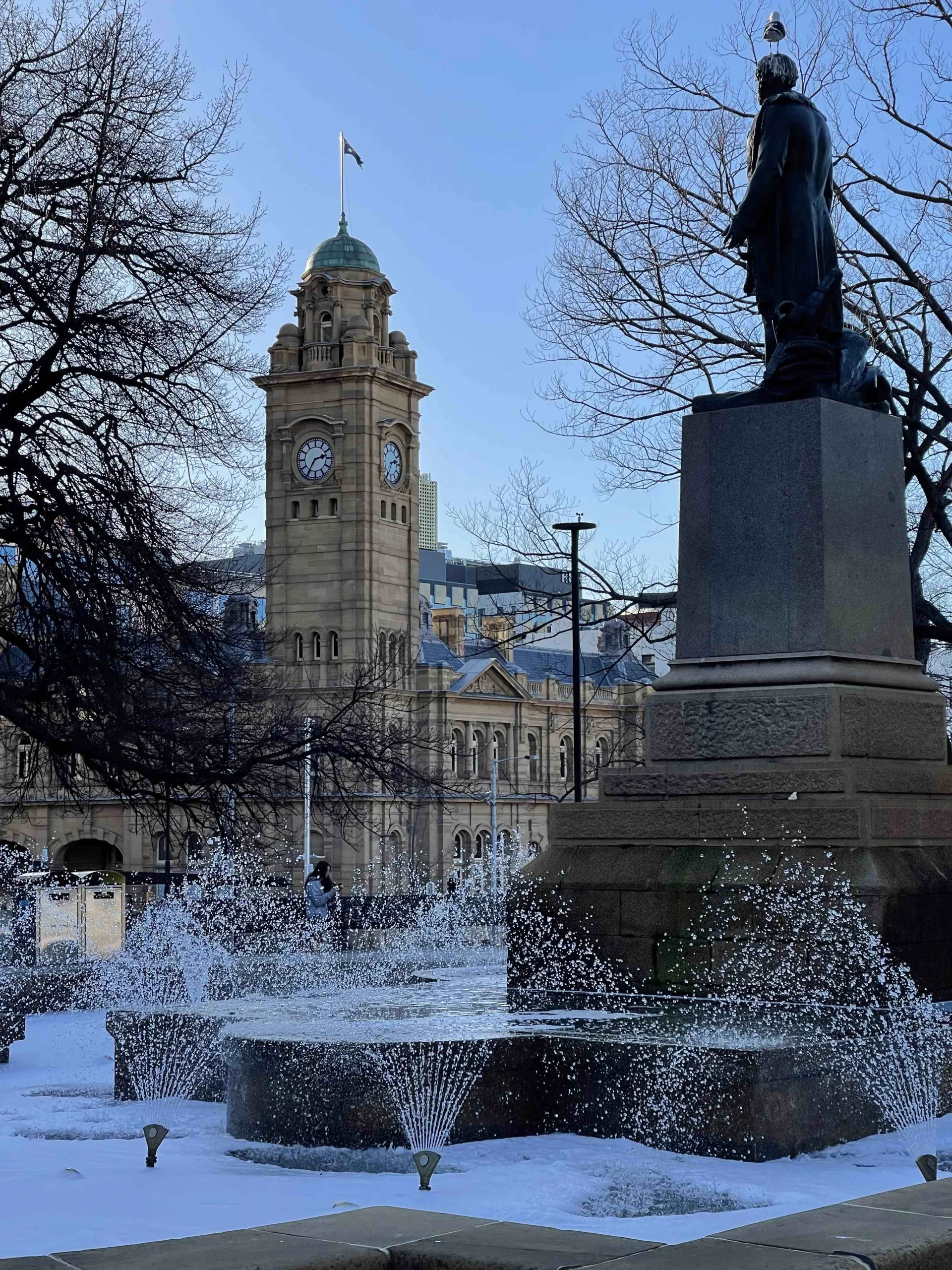 A view of Franklin Square in Hobart, featuring the historic clock tower of the Post Office and statue framed by bare trees. In the foreground, a fountain sprays water upward, catching the light.
