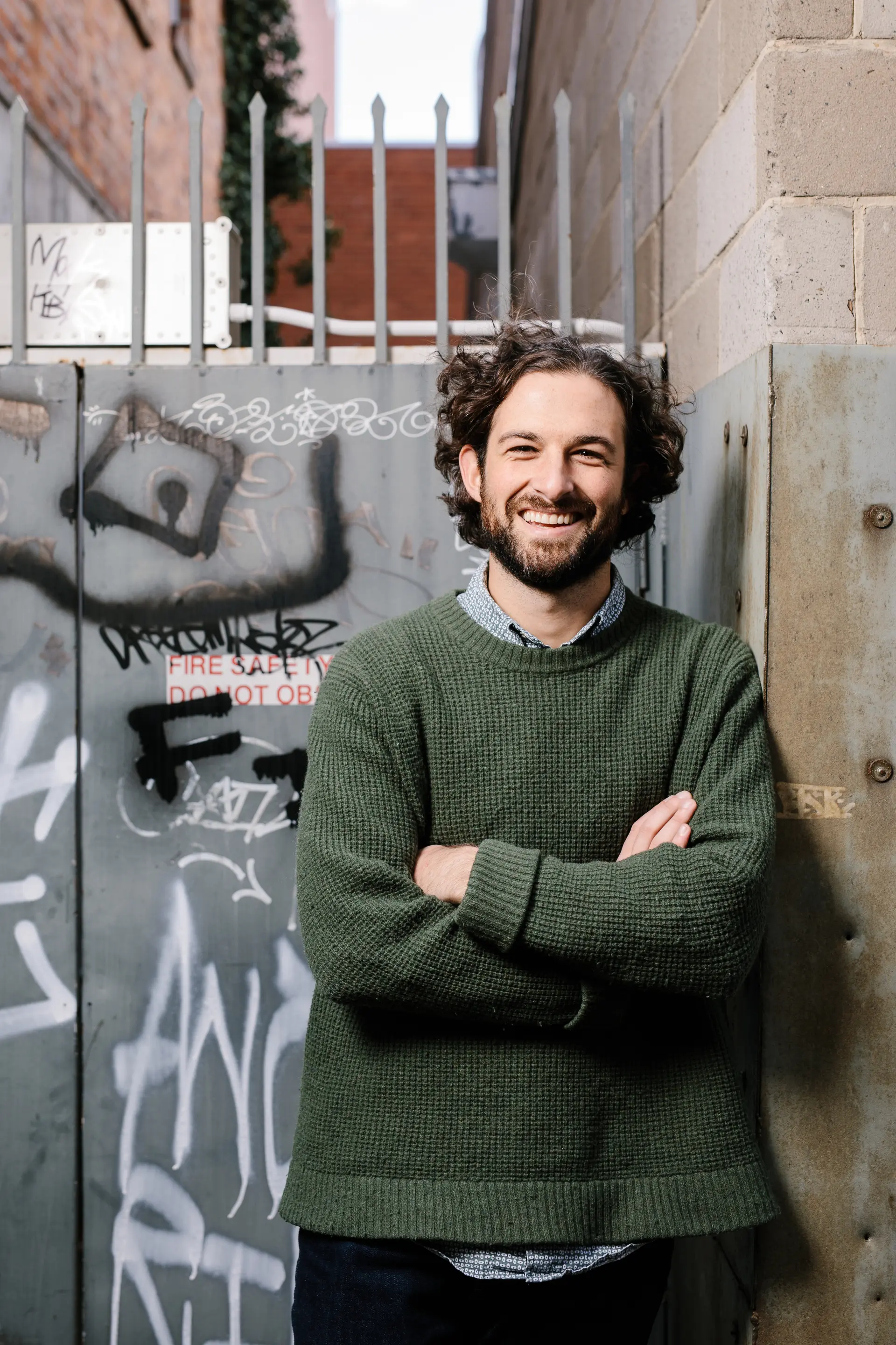 A professional headshot of Tim Blakeway standing in an urban setting against a graffiti-marked metal gate and concrete wall. He wears a deep green knit sweater over a collared blue shirt, with curly dark brown hair and a short beard, arms folded and smiling directly at the camera with a relaxed and friendly expression.
