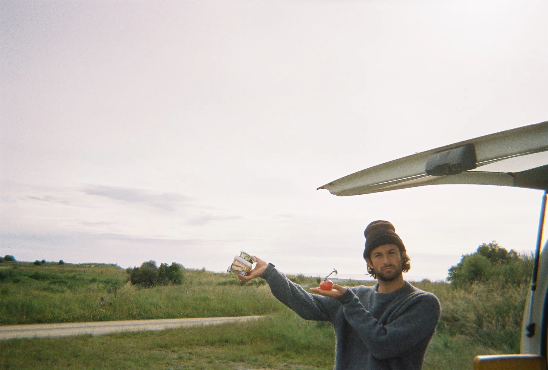 A candid photo of Tim outdoors beside an open car boot in a grassy rural landscape, wearing a beanie and holding up a small tomato and a dips with a playful expression.