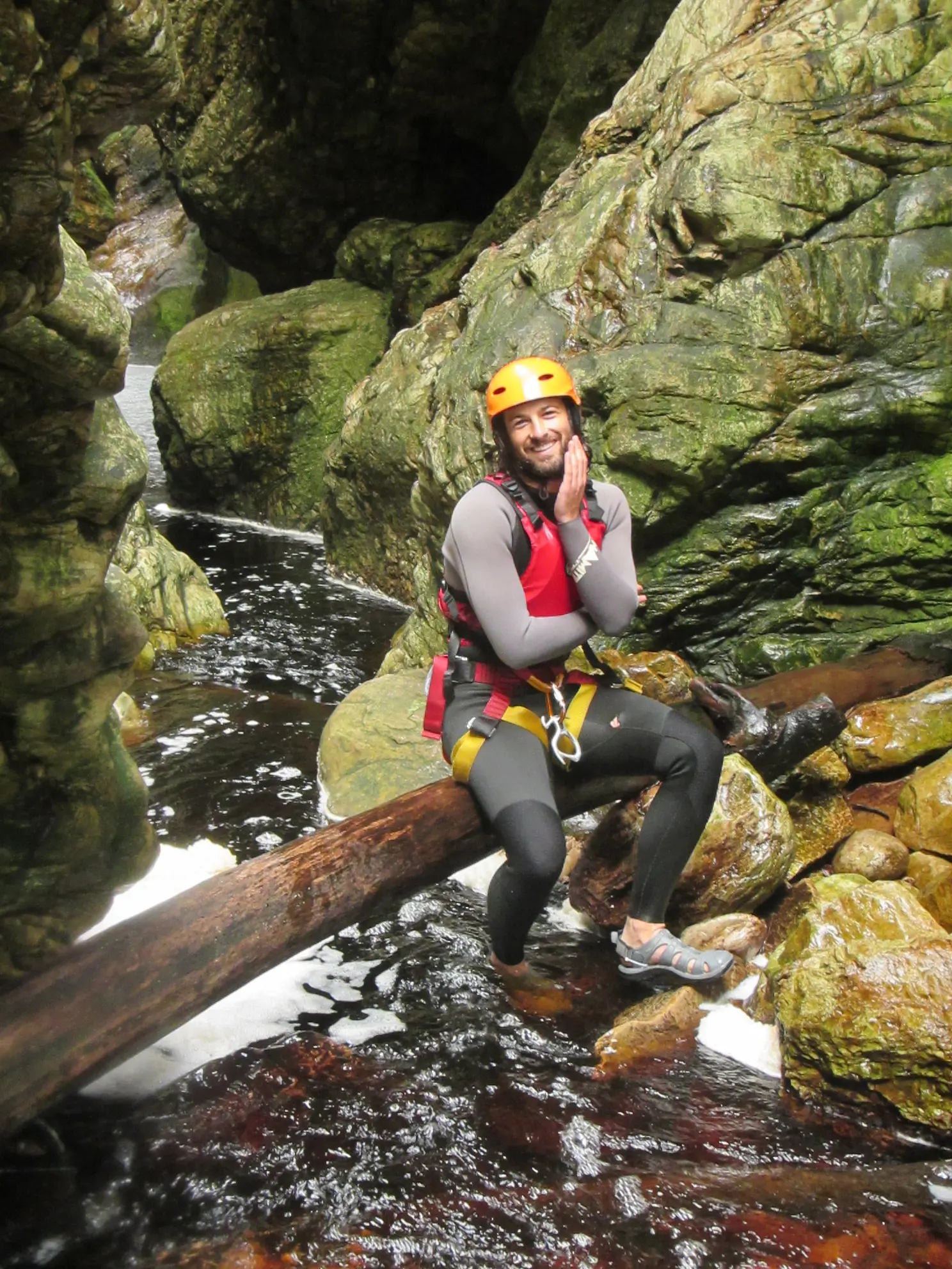 Tim sits on a log above a narrow stream within a rocky canyon, wearing a helmet and canyoning gear, smiling with one hand resting on his face playfully as water flows below.