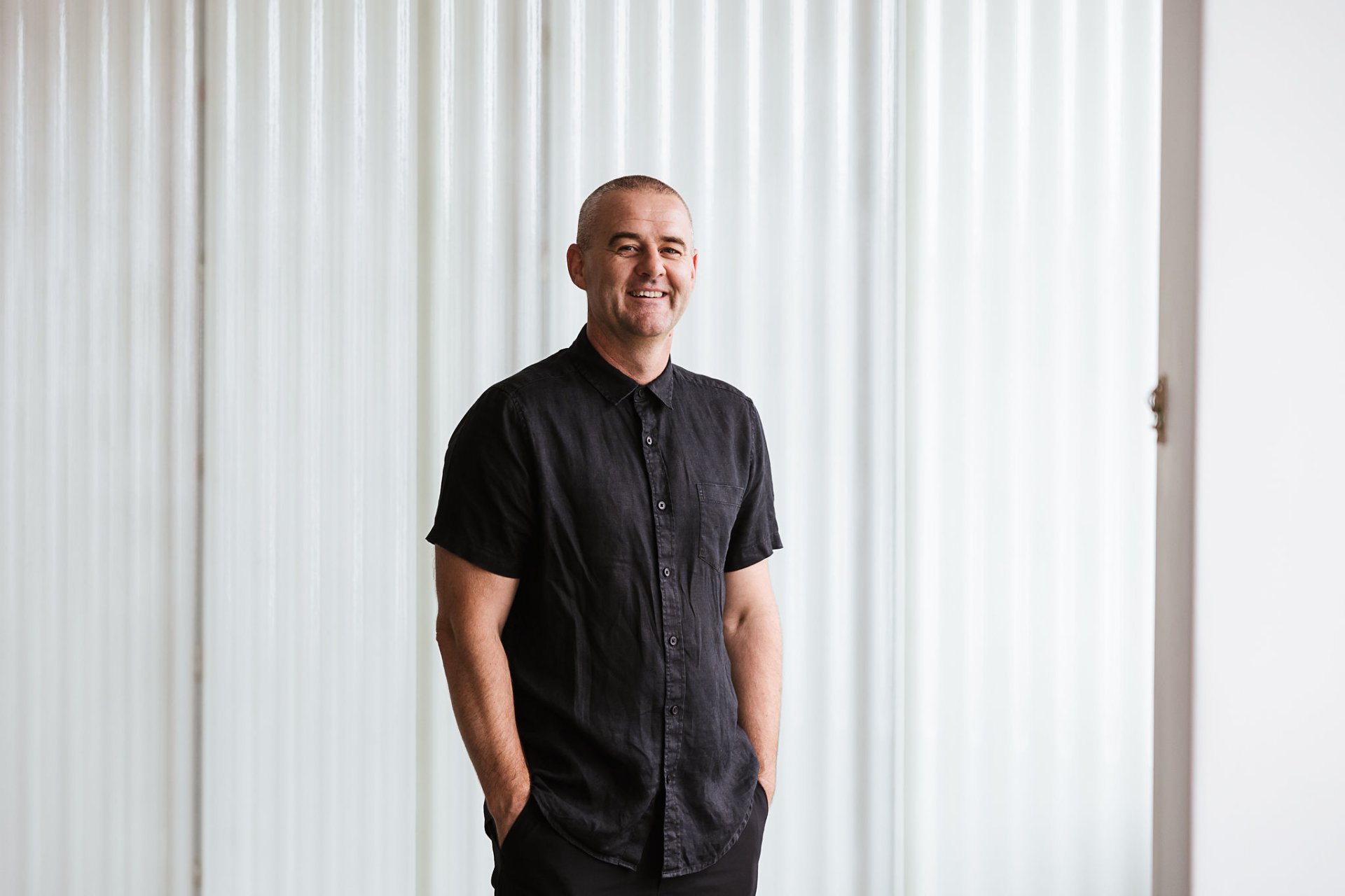 A professional headshot of Todd Henderson standing against a light polycarb backdrop in our Hobart Studio. He wears a black short-sleeve button-up shirt, smiling directly at the camera with a relaxed and approachable expression, hands casually in his pockets.