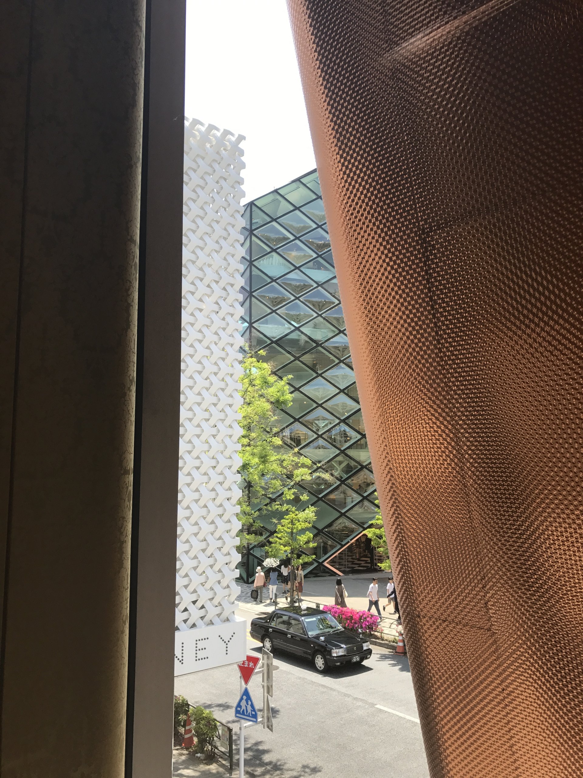 Looking out of a building in Tokyo towards a Japanese street framed between textured architectural elements, geometric glass building and patterned facade, with pedestrians, a parked car, and greenery in the foreground.